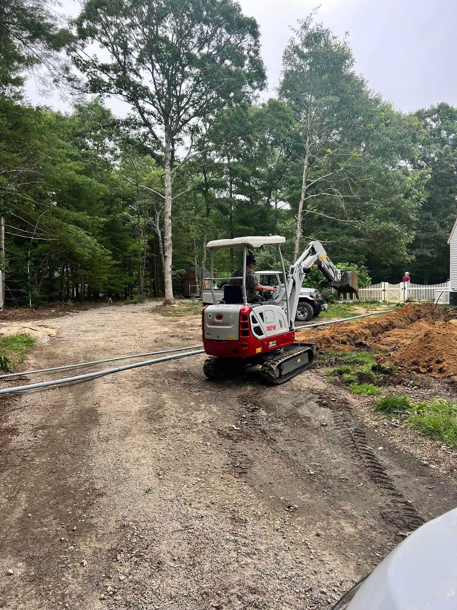 Red and white mini excavator on a gravel path beside trees and a wooden fence, clearing soil and debris.