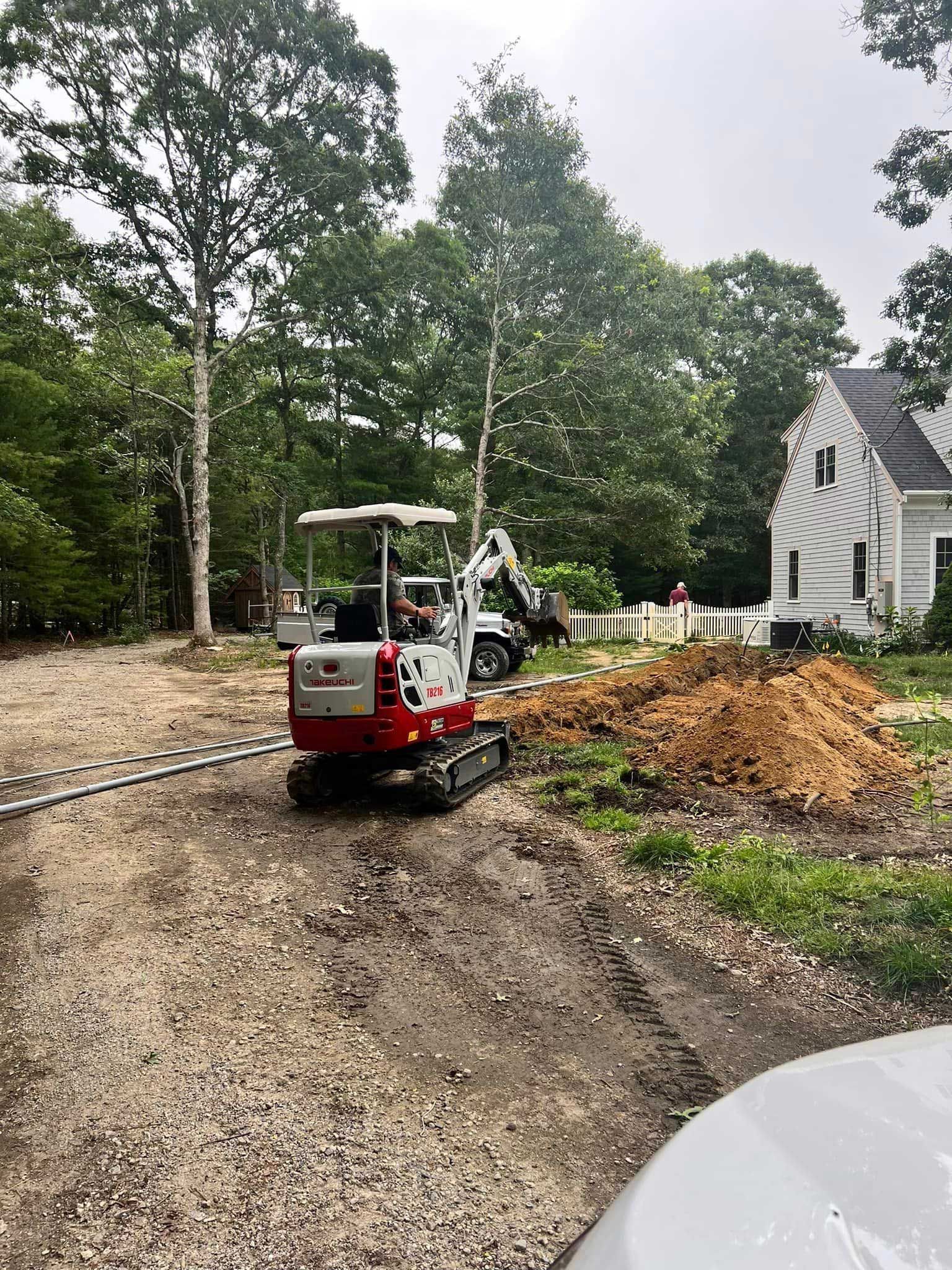 Small red excavator on a gravel driveway beside a house, moving a dirt pile near trees and a fence.