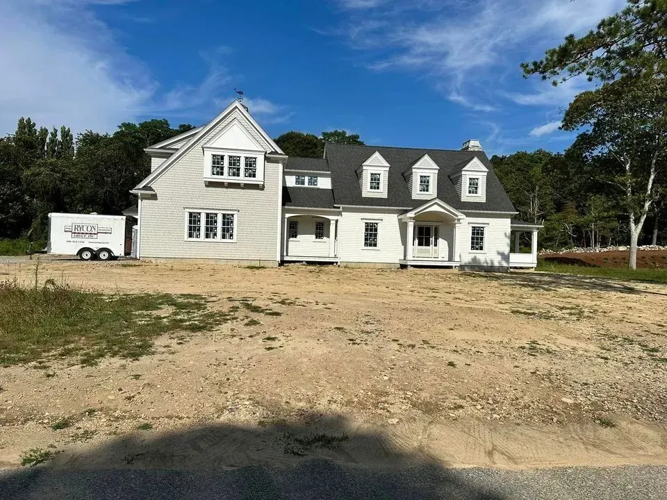 Large gray house under construction on a dirt lot with a white truck nearby and trees in the background