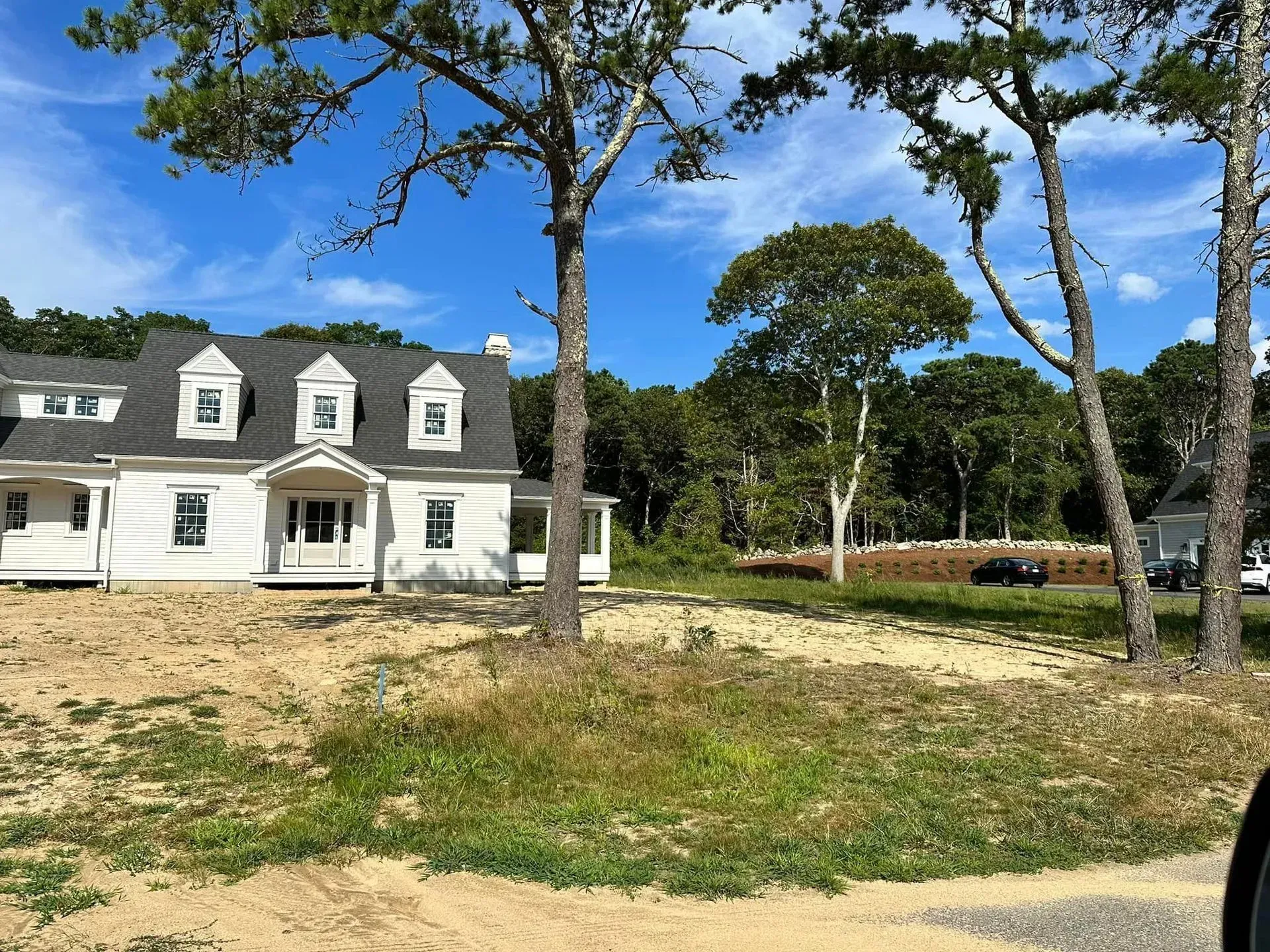 White house with dormer windows on a sandy, tree-lined lot under a bright blue sky