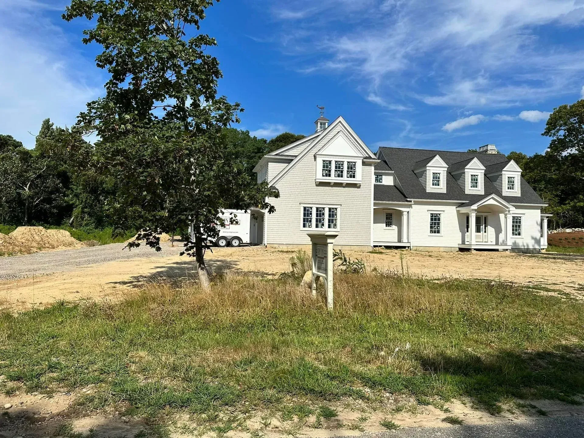White house with gray roof and front yard, beside a large tree under a blue sky.