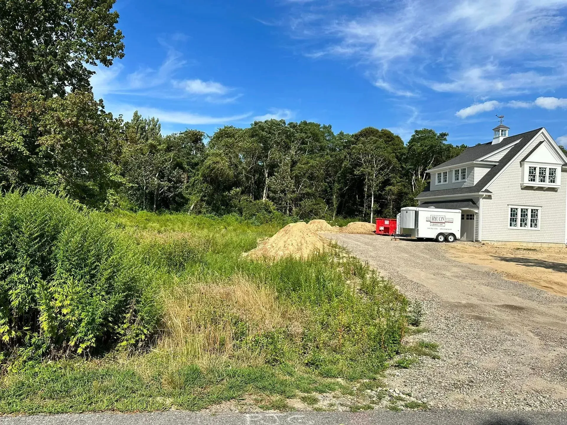 Gravel driveway beside a white house, with a sand pile and trees under a blue sky.