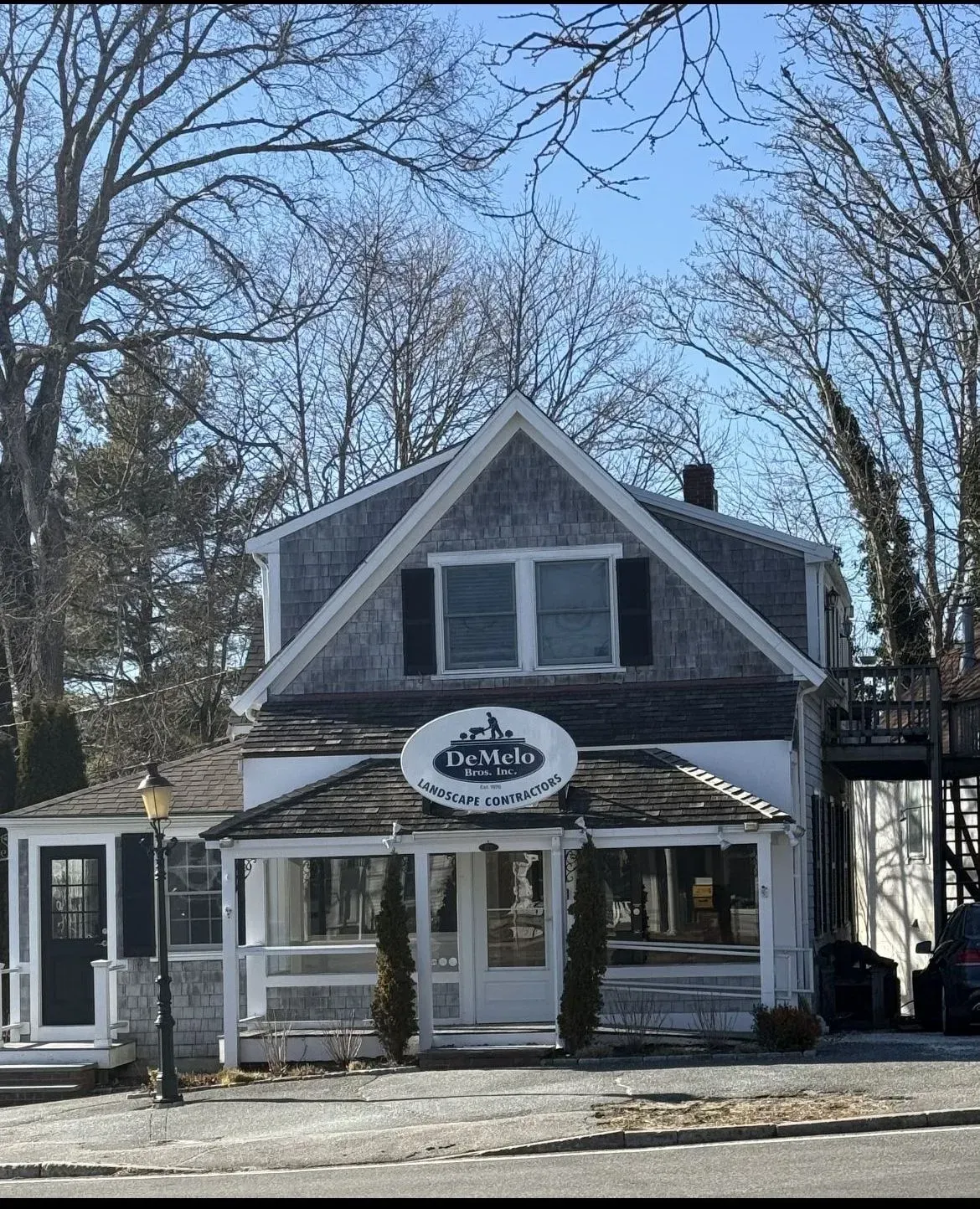 Small gray storefront with white trim and a round sign, framed by leafless trees under a blue sky