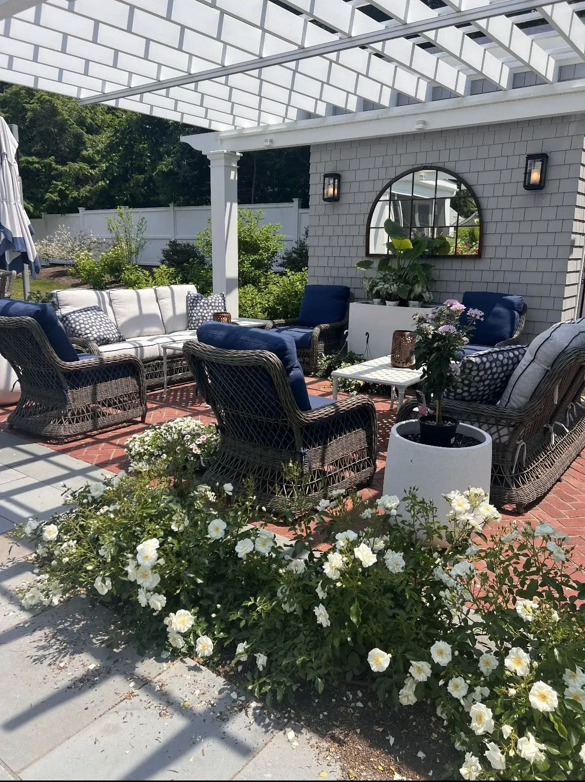 Outdoor patio with wicker chairs, cushioned seating, and white flowers under a pergola beside a stone wall