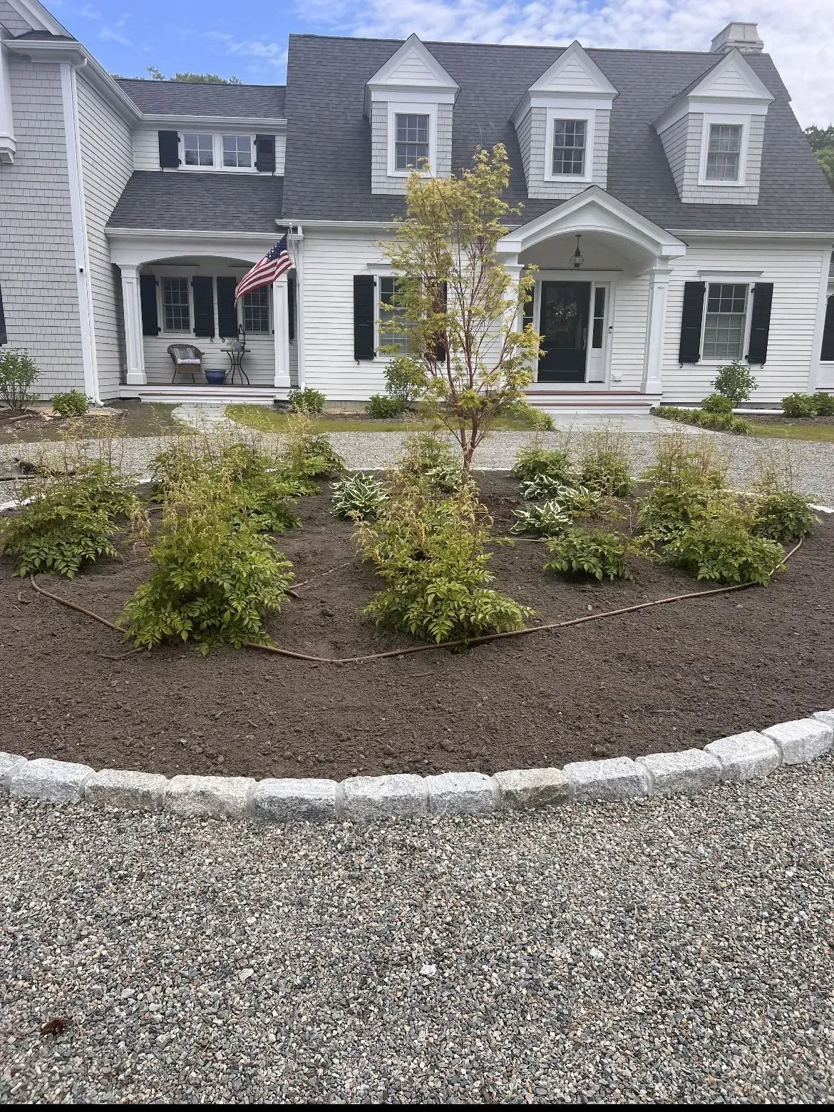 White house with gray roof and dormers, front shrubs, and a gravel driveway in the foreground.
