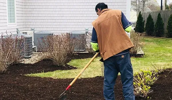 Person using a rake to spread mulch in a garden bed beside a house