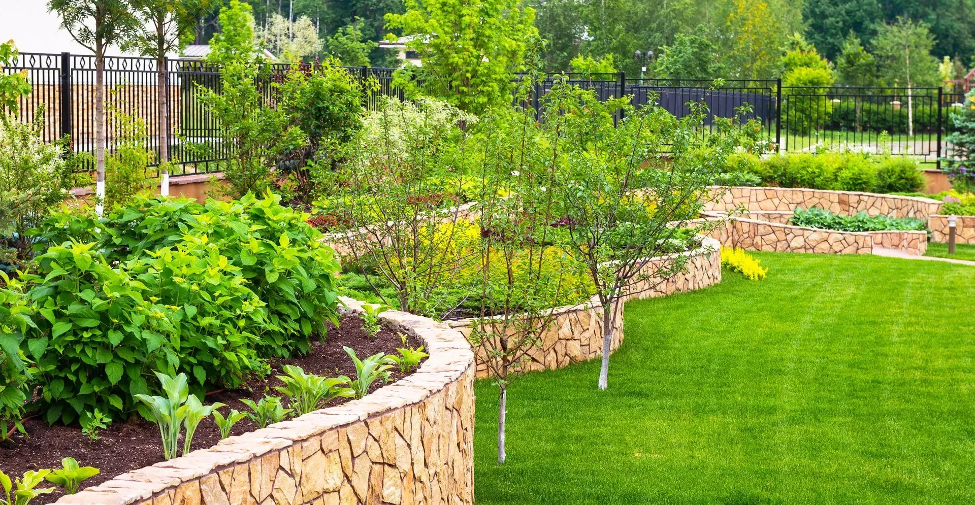 Curved stone garden beds with lush green shrubs beside a neatly trimmed lawn and trees