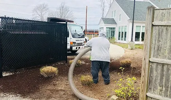 Worker vacuuming mulch beside a fence and house, with a truck parked nearby.