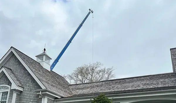 Blue crane lifting materials over a gray house roof under an overcast sky