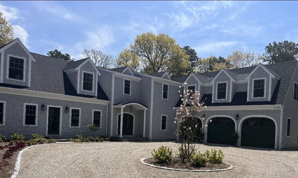 Large gray house with dormer windows, arched garage doors, and a gravel driveway under a clear sky
