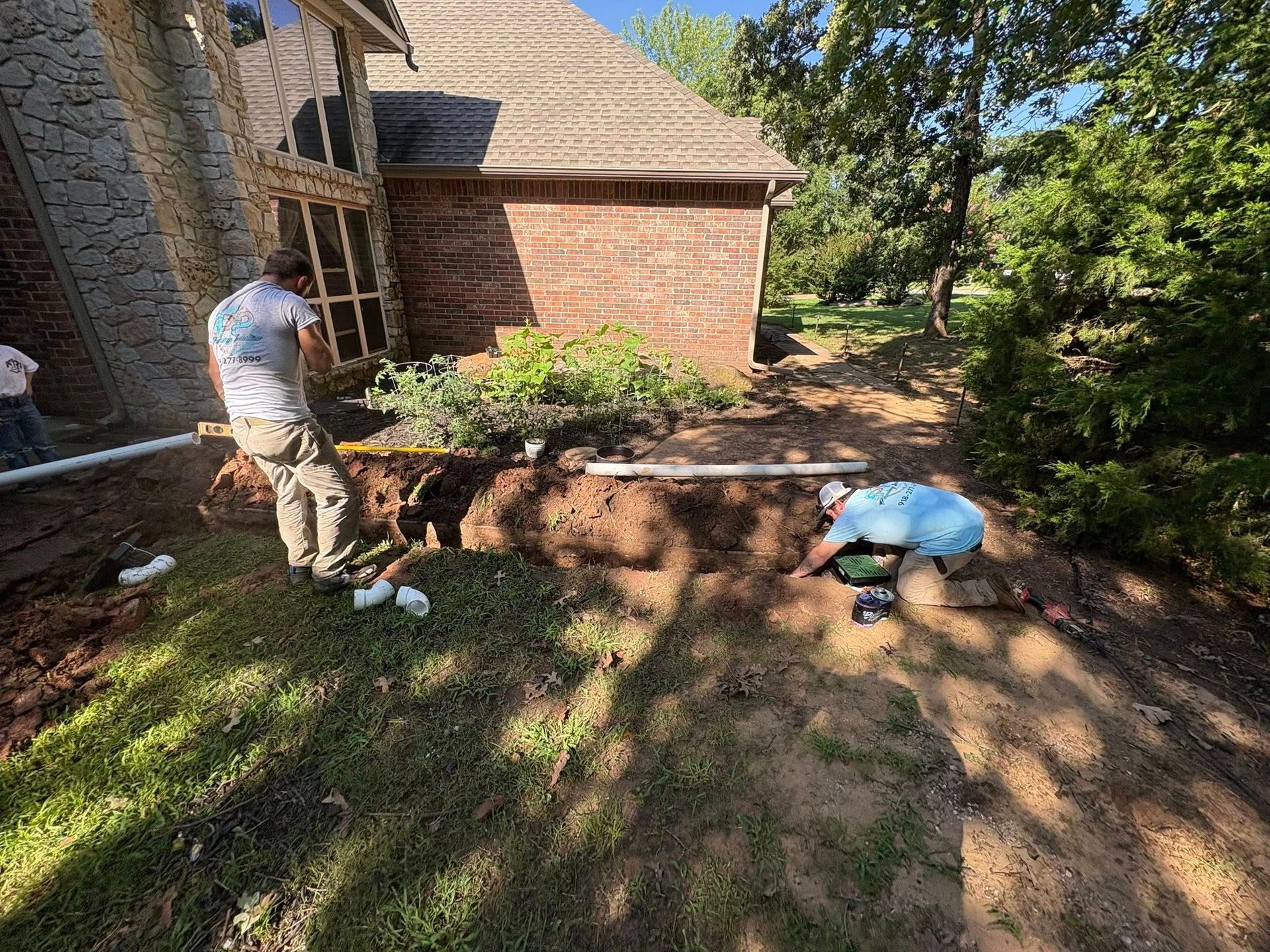 Two workers install drainage pipes in a trench near a brick building with a sloped roof and lush greenery.
