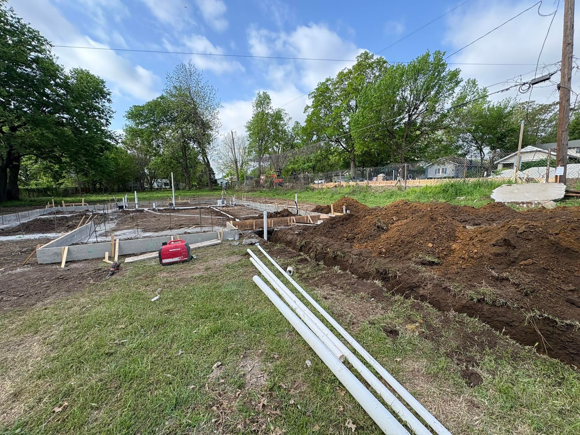Construction site with concrete forms, piping, and dirt mounds on grass under a cloudy sky.