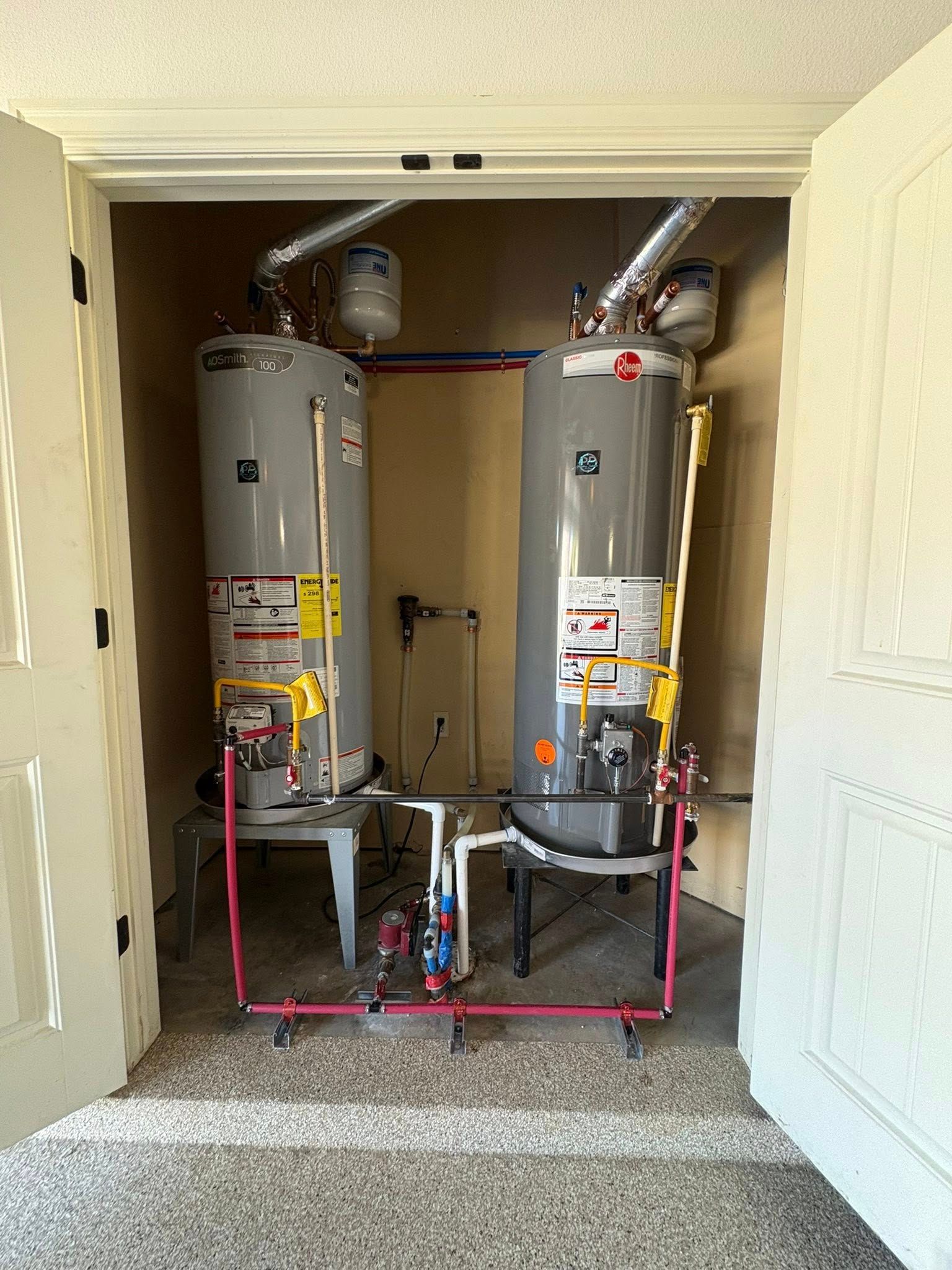 Two water heaters inside a doorway with pink and metal pipes.