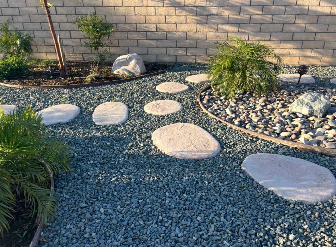 A stone path through a garden bed with blue gravel and stepping stones, bordered by a brick wall and plants.