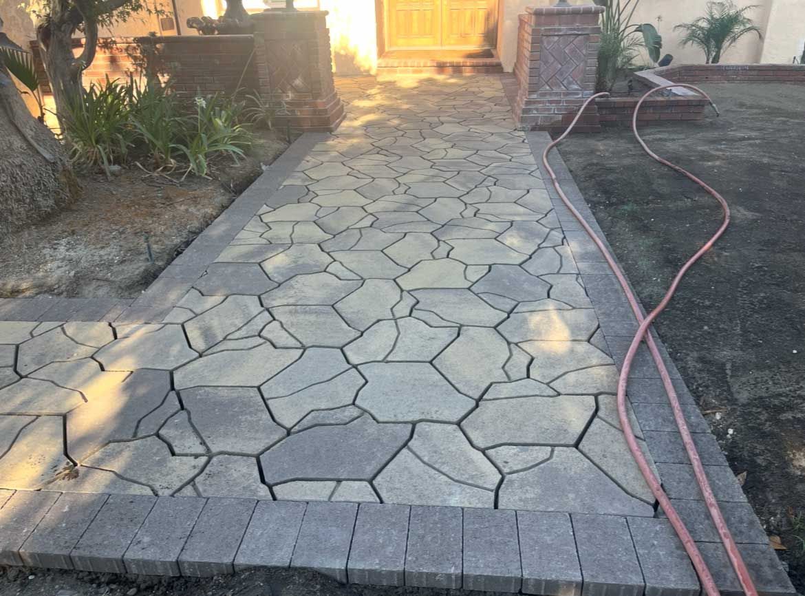 Stone walkway with dark border leads to a house, a hose is lying on the grass.