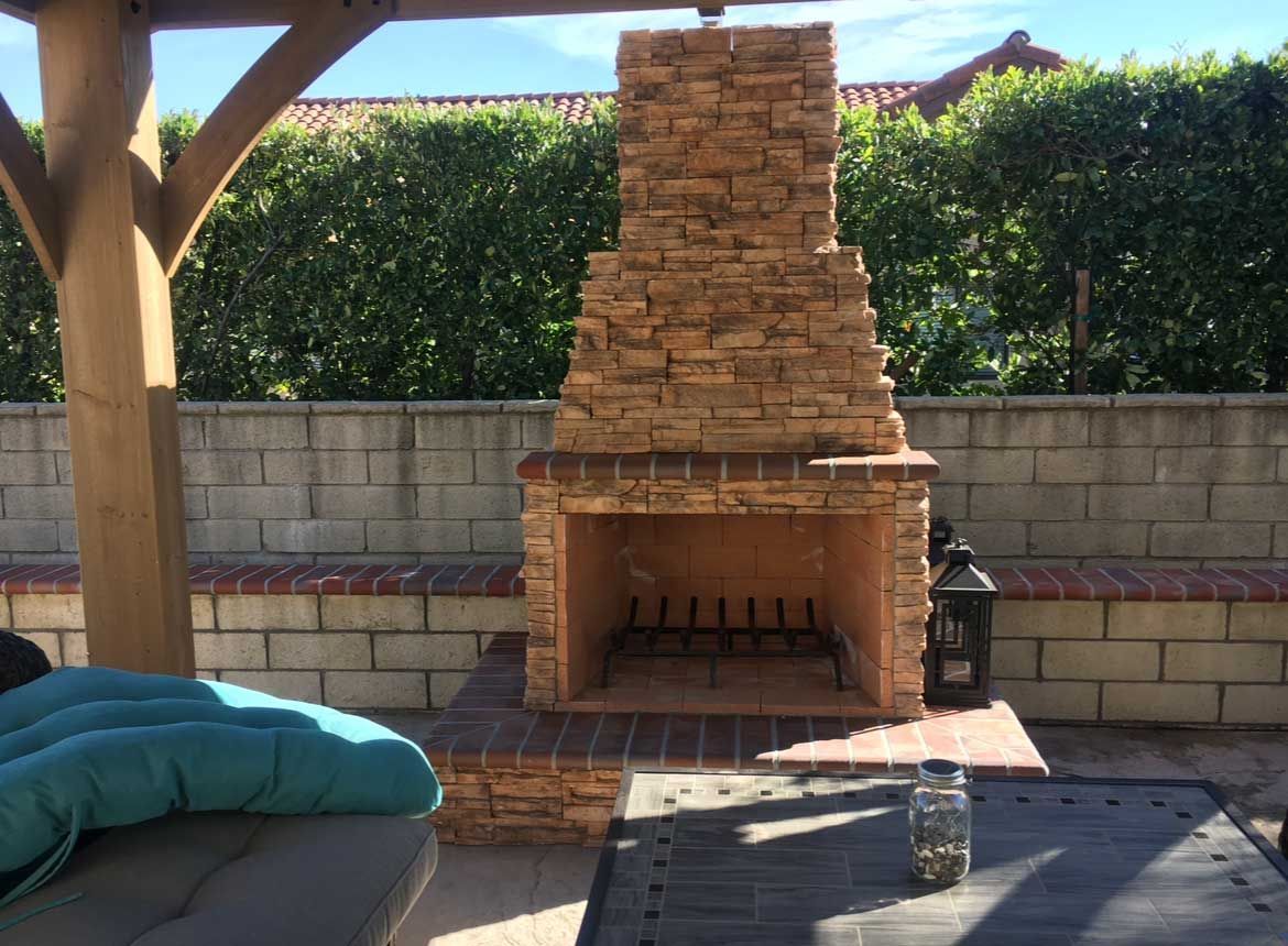 Outdoor brick fireplace in a patio setting; with seating, and a green hedge background.