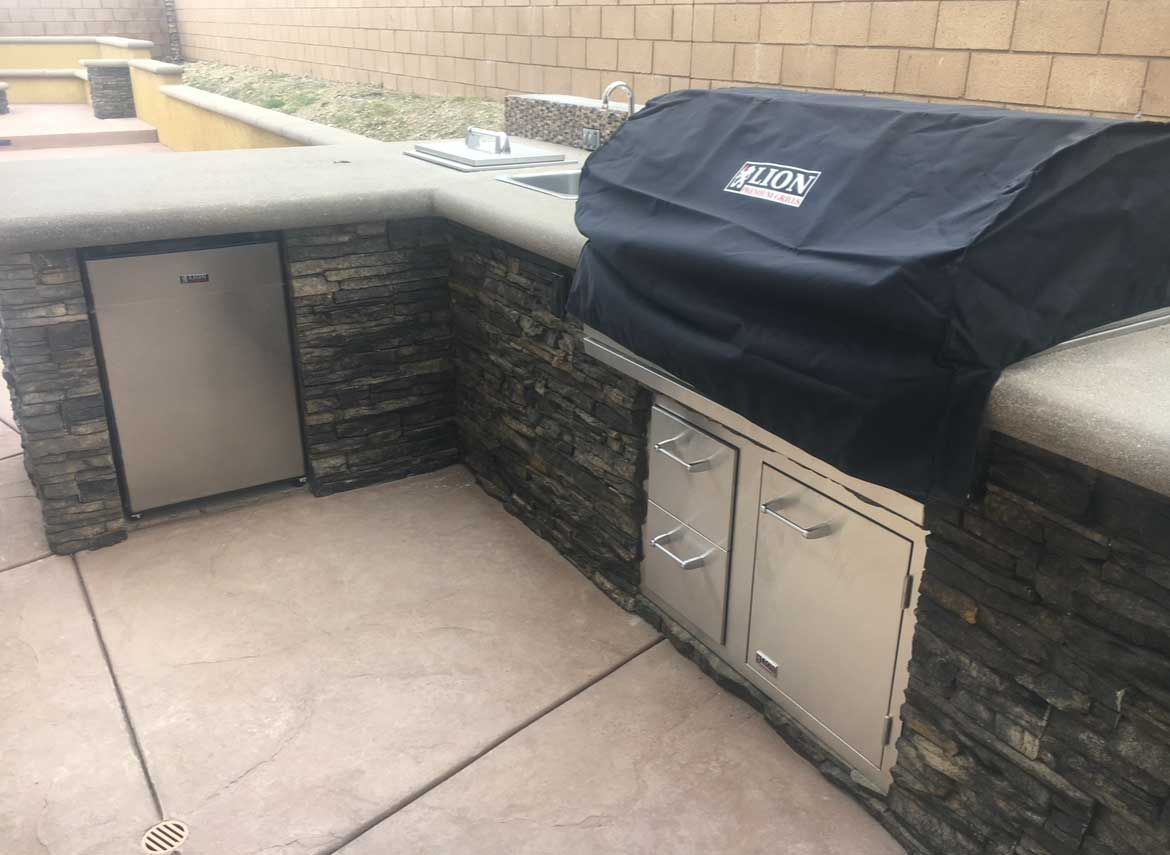 Outdoor kitchen with stainless steel appliances, stone facade, and grill covered with a black lid.