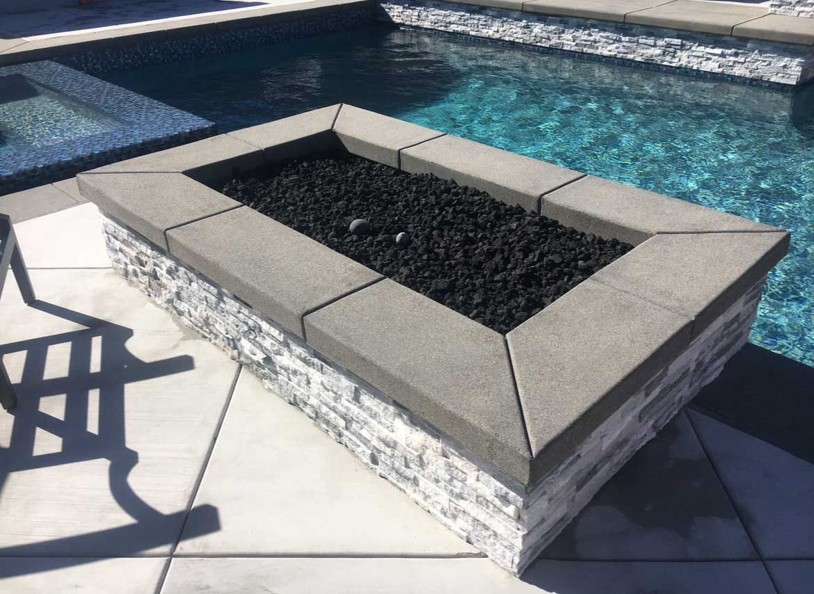 Fire pit with black lava rocks by a pool. Stone base and concrete border.