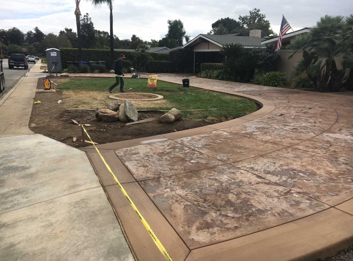 Stamped concrete driveway under construction, with a worker, rocks, and grass.