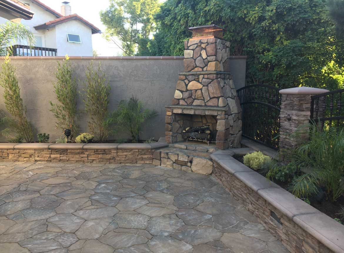 Stone outdoor fireplace with a paved patio surrounded by plants and walls.