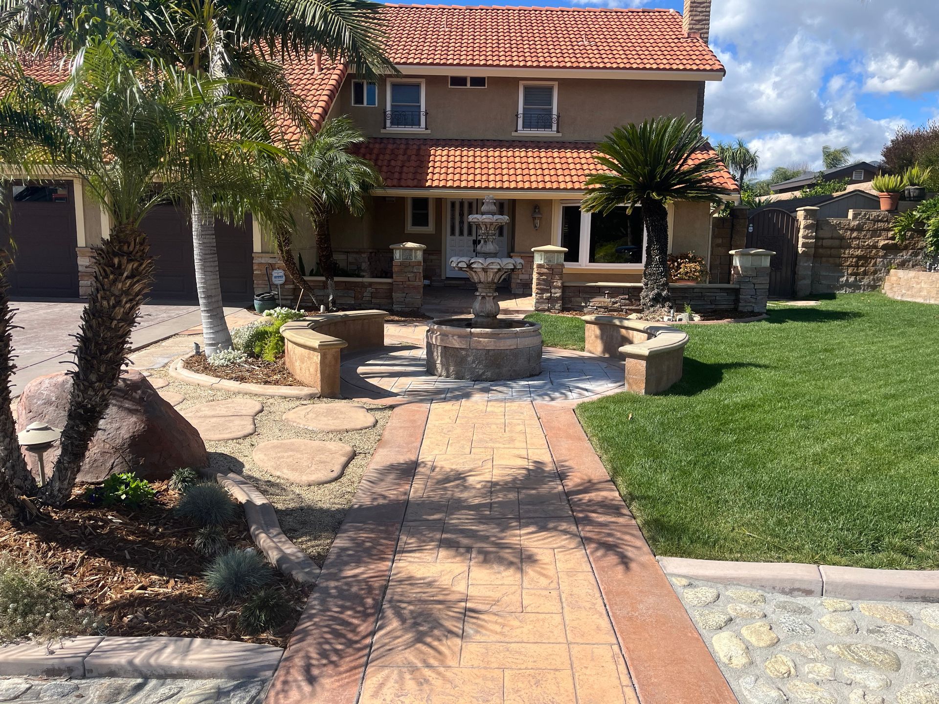 House with tile roof, fountain, and palm trees in front yard. Paved walkway. Green lawn.