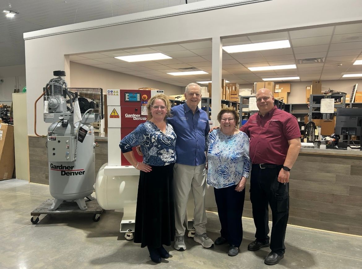 Four people stand in front of an air compressor inside a shop.