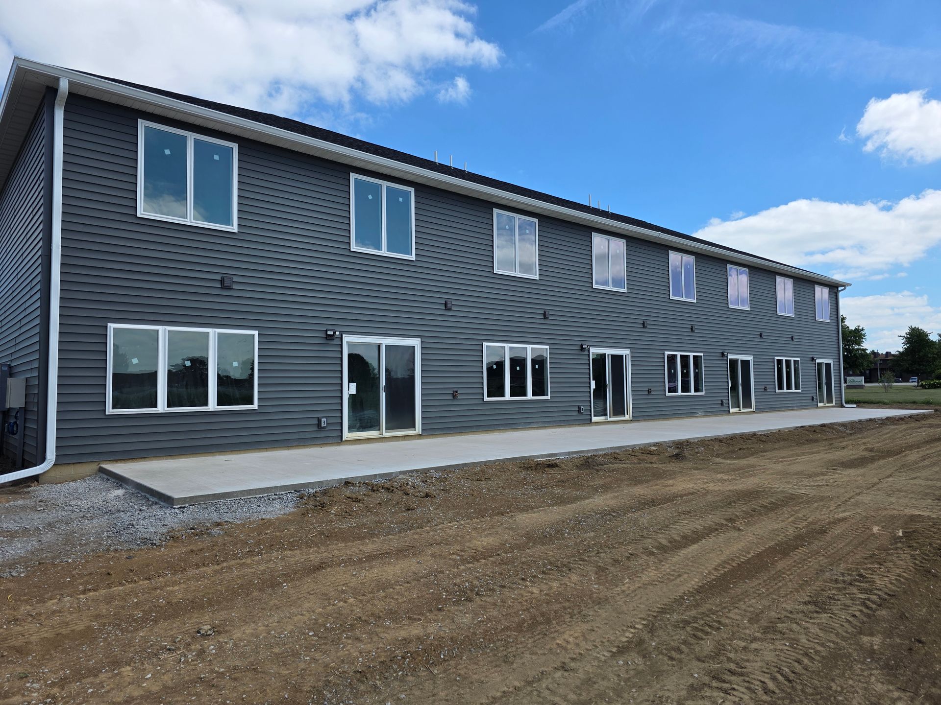 Dark gray apartment building exterior with large windows, concrete patio, and dirt ground.