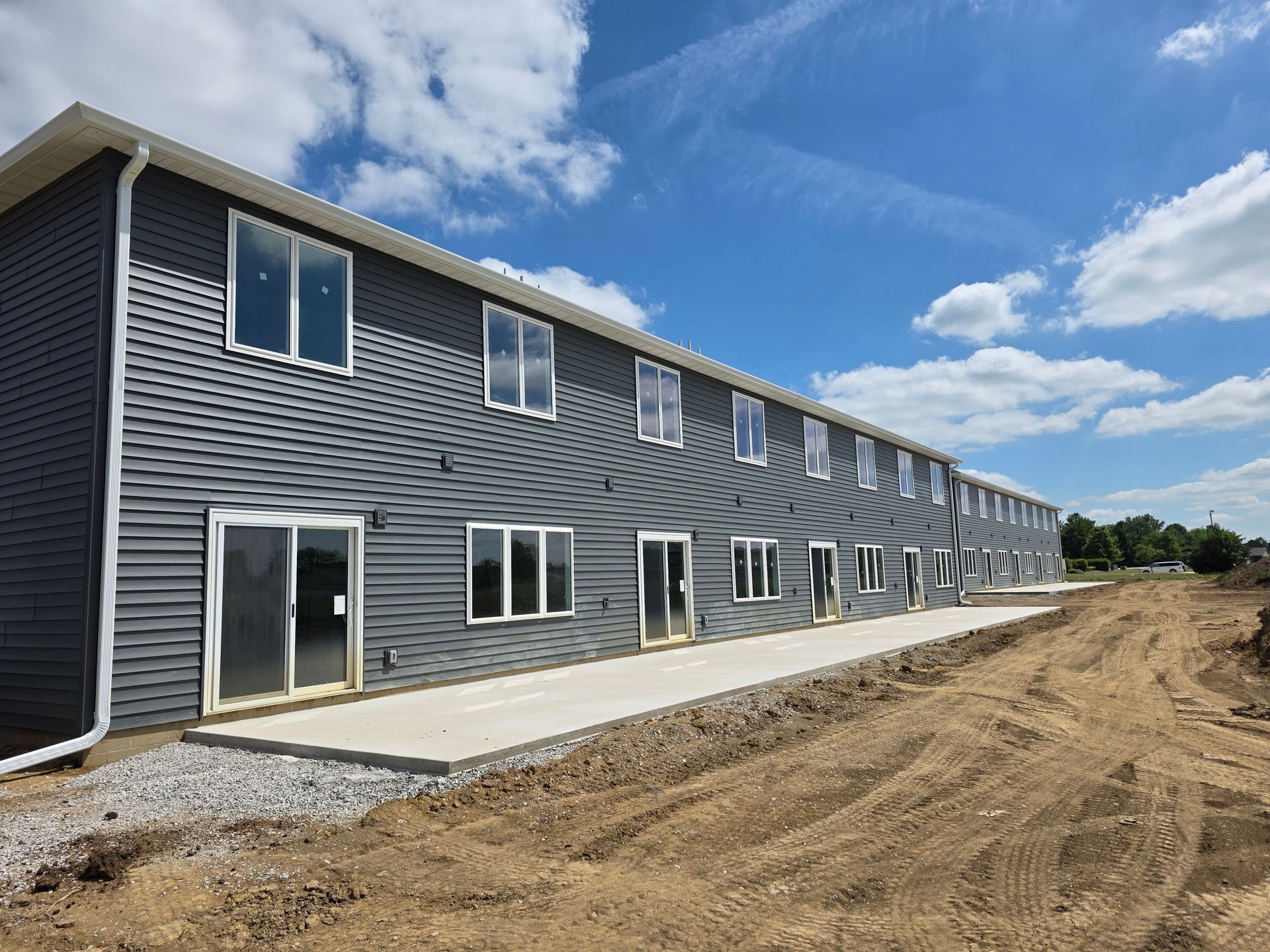 Two-story gray building with multiple windows and sliding doors, concrete patio, dirt, and blue sky.