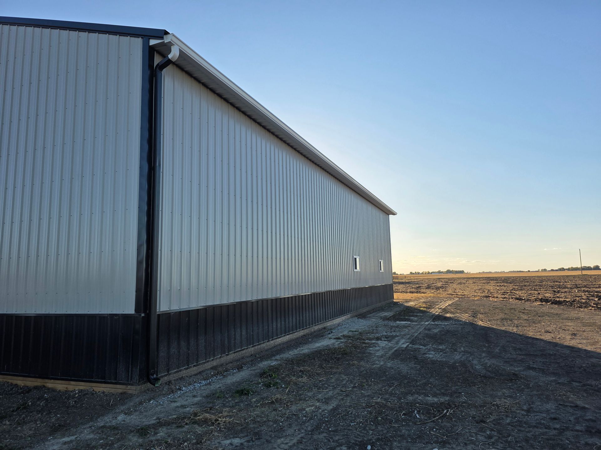 A long, white and black metal-sided building on a gravel lot under a bright blue sky.