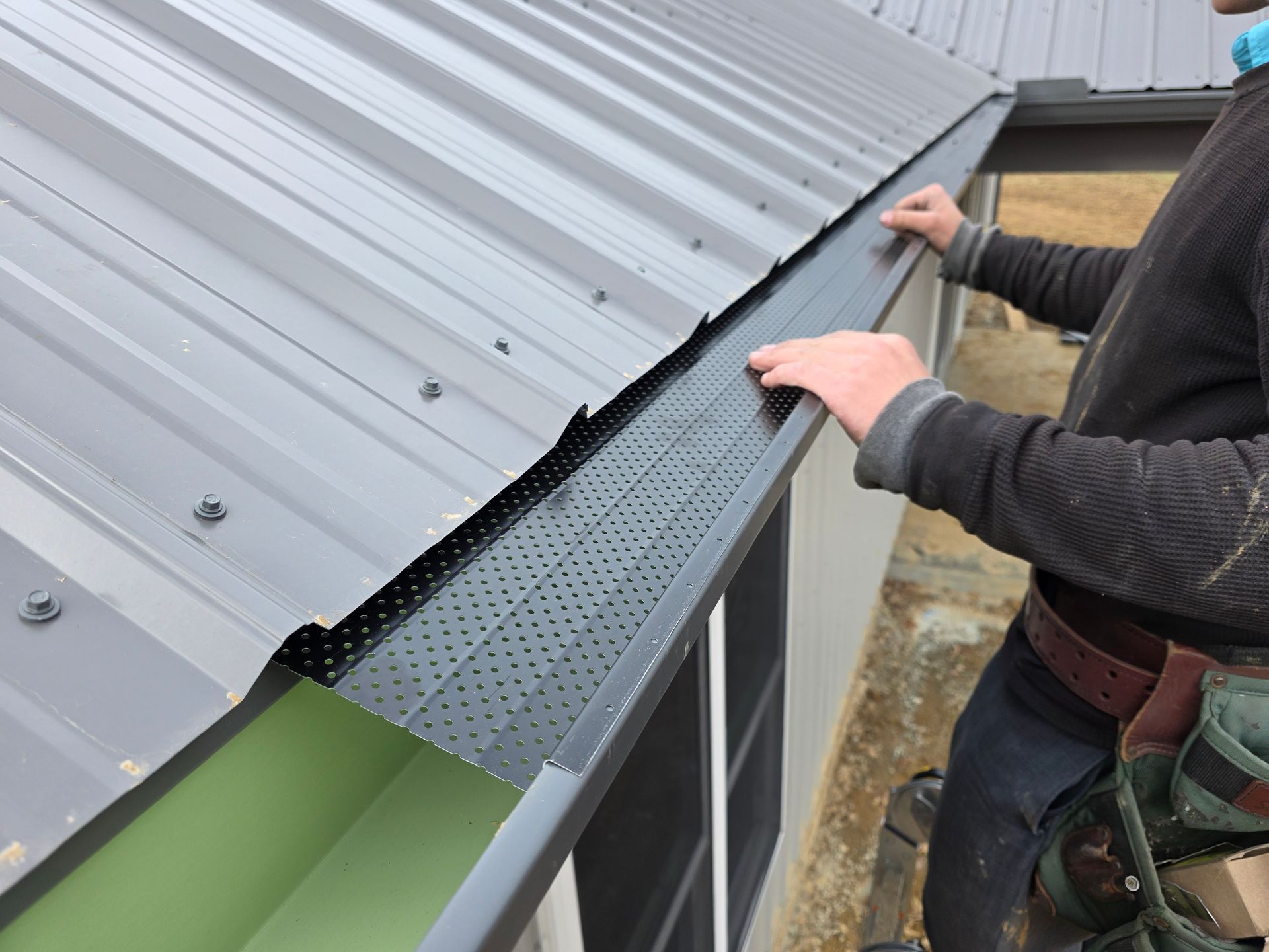 Person installing a black perforated gutter guard on a metal roof.