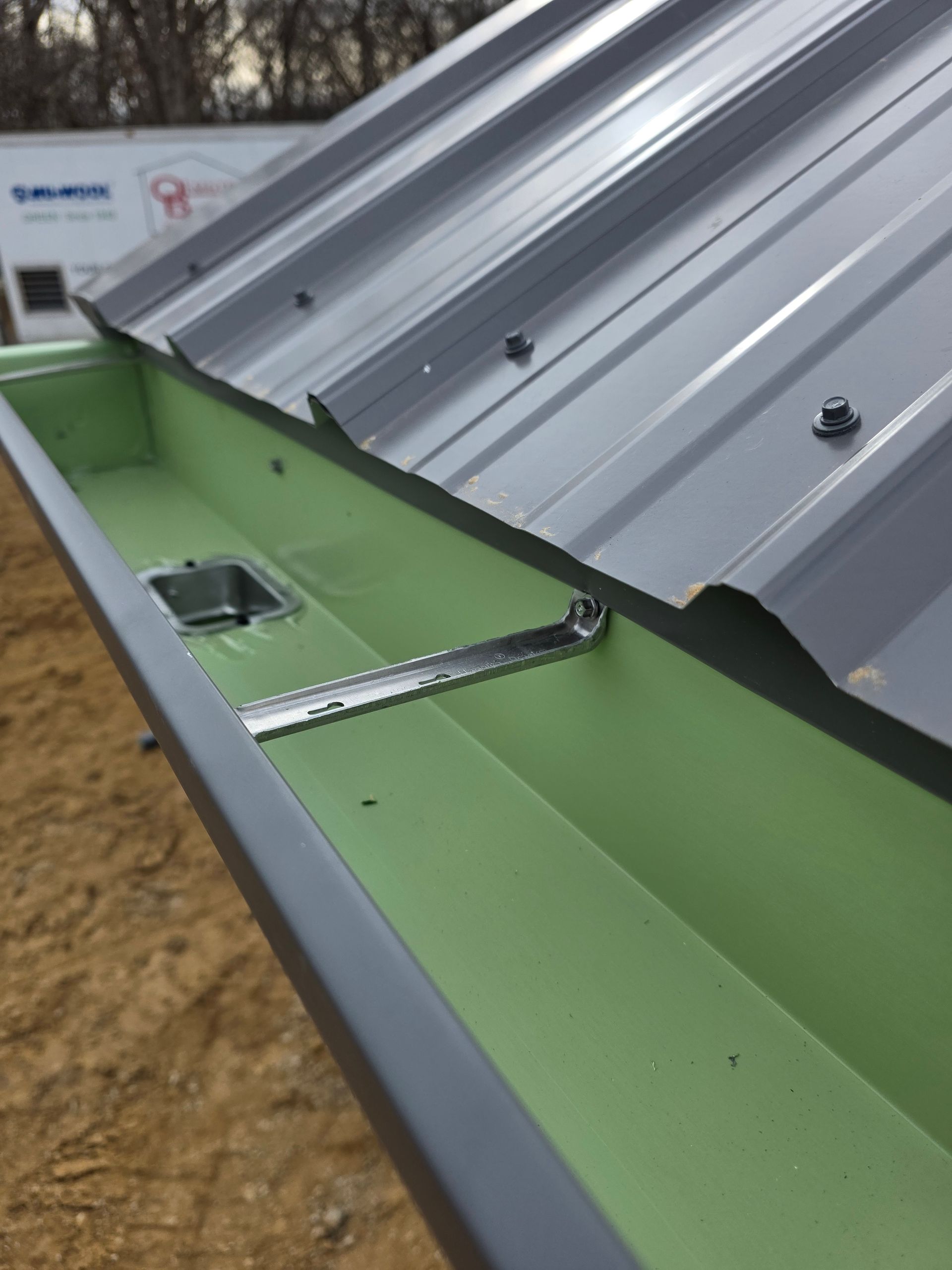 A metal roof gutter with a green interior, attached to a corrugated metal roof. A metal bracket secures the gutter.