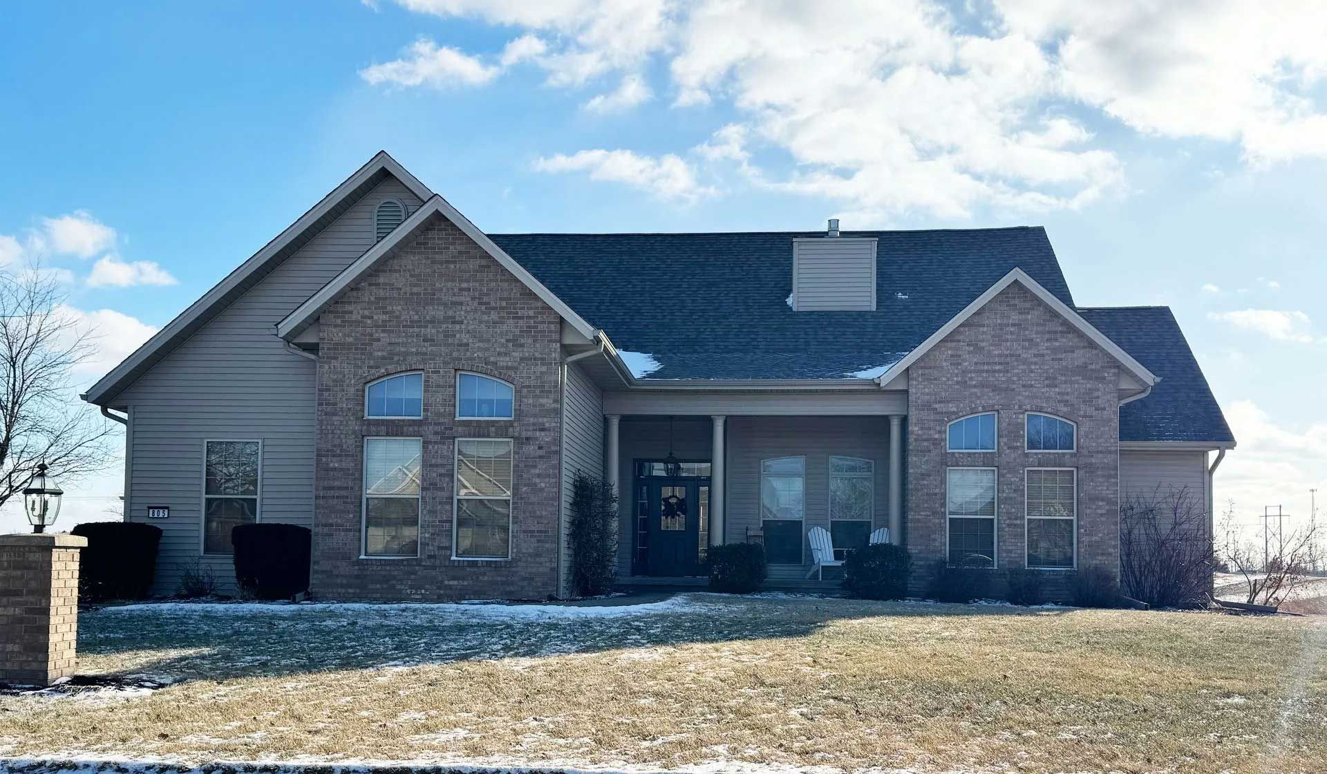 Tan brick and siding house with a porch and snow on the roof under a partly cloudy sky.