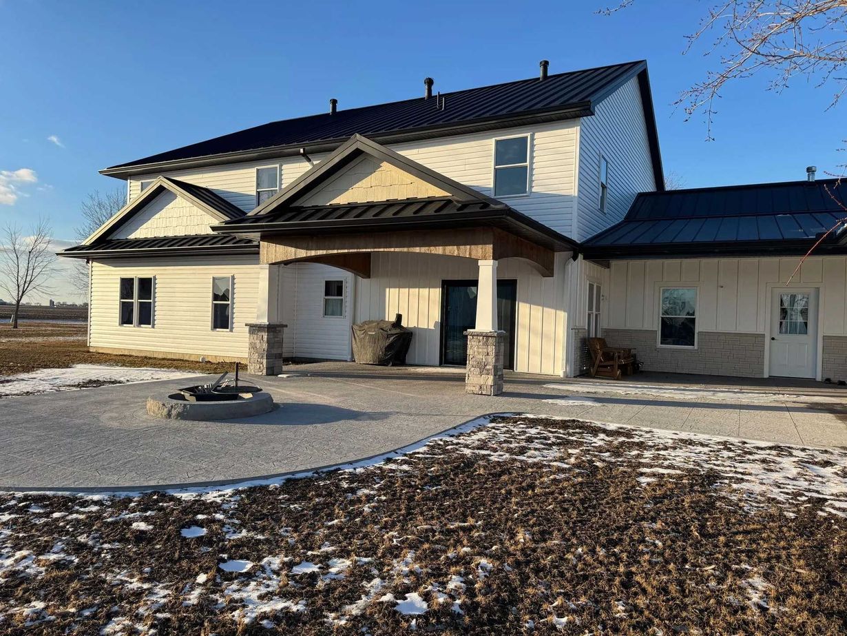 Two-story white house with black roof, covered porch, and stone accents. Blue sky and gravel patio in foreground.