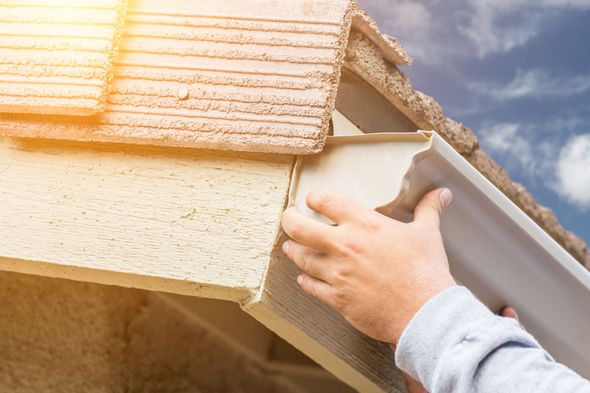 Person installing a section of light-colored gutter on a beige house, with a blue sky background.