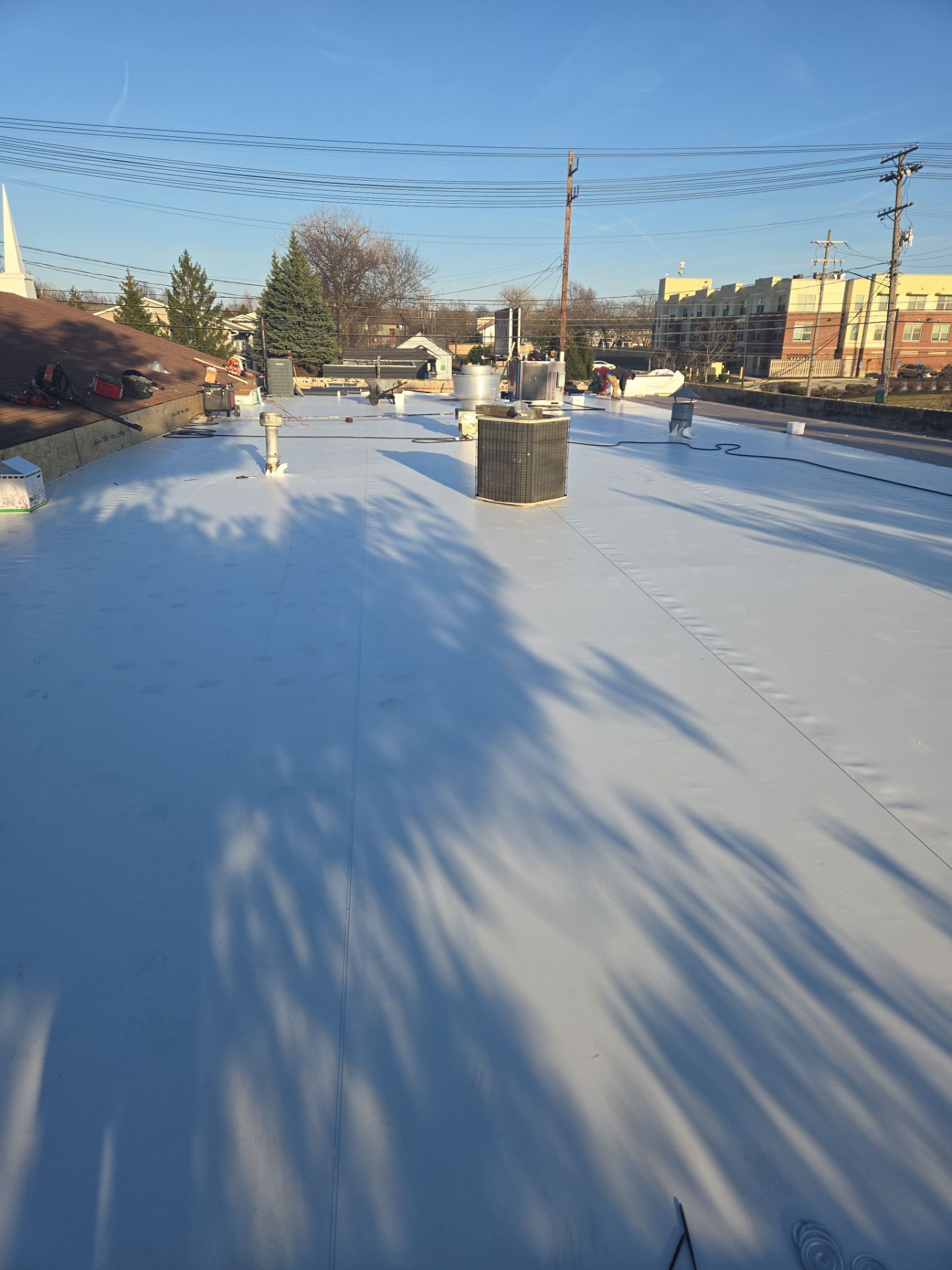 White commercial roof under a clear blue sky; shadows of trees and utility poles are cast upon the surface.