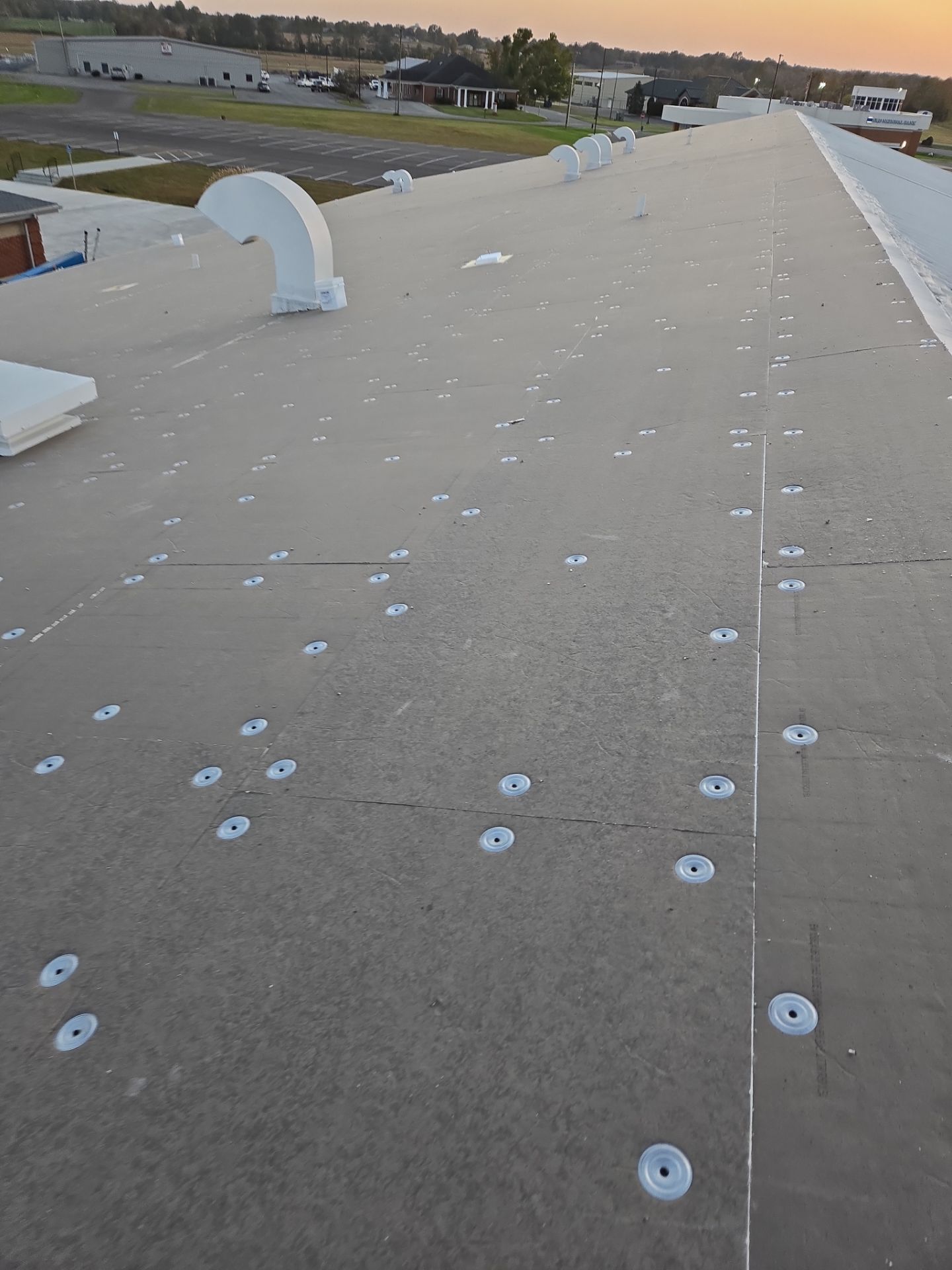 Flat commercial roof with numerous fasteners, white vent, and buildings in the distance.