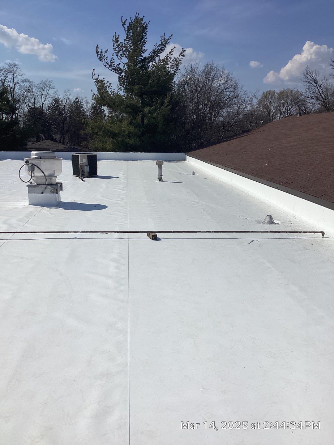 White flat roof with vents and a brown shingled roof in the background under a blue sky.