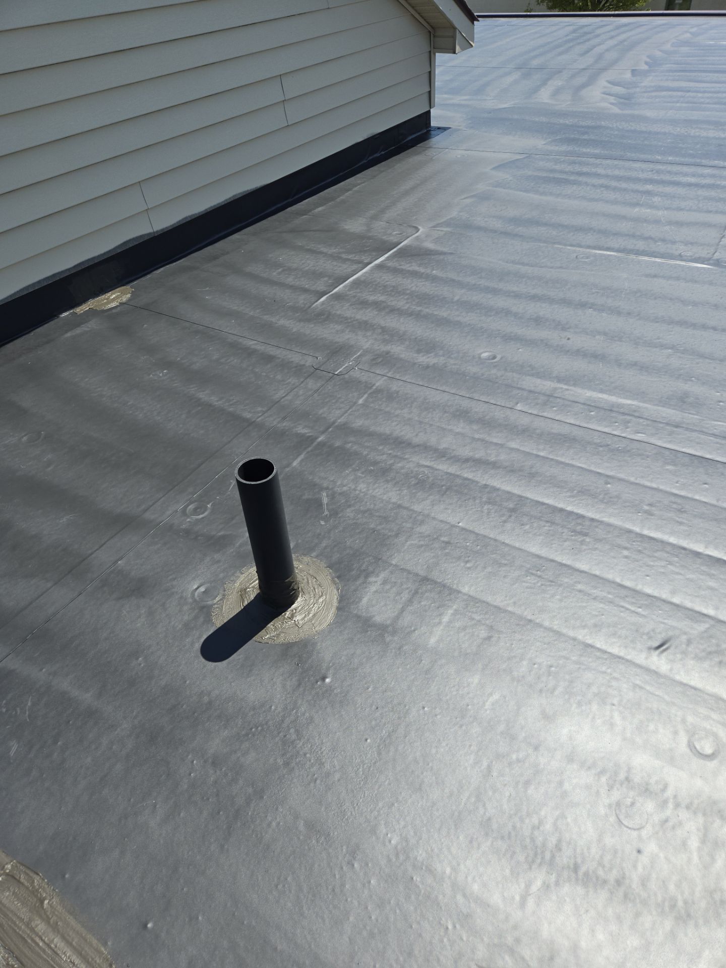 Flat black rooftop with a black pipe; a wall of white siding on the side.