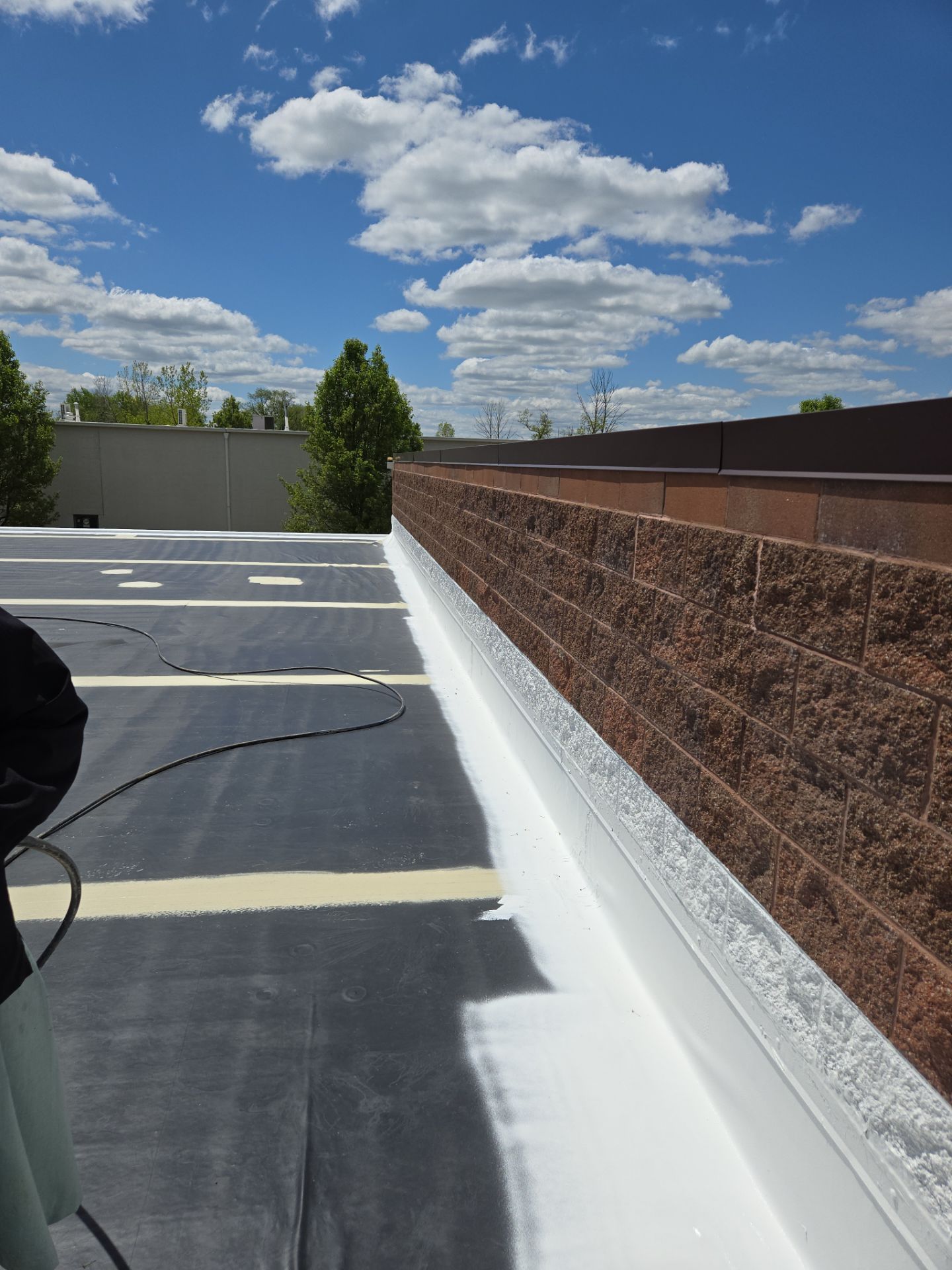 A flat roof with white sealant along a brick wall under a blue sky with clouds.