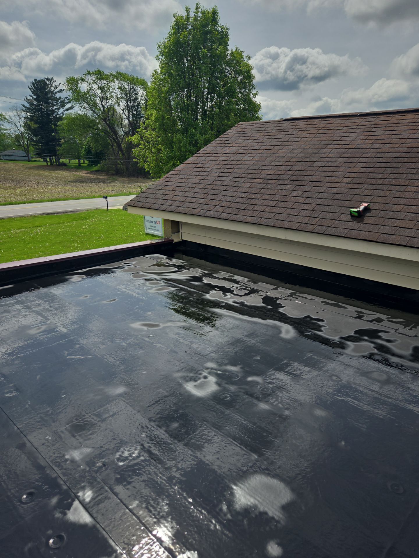 Flat black roof with water puddles, brown shingled roof in background, sunny sky.