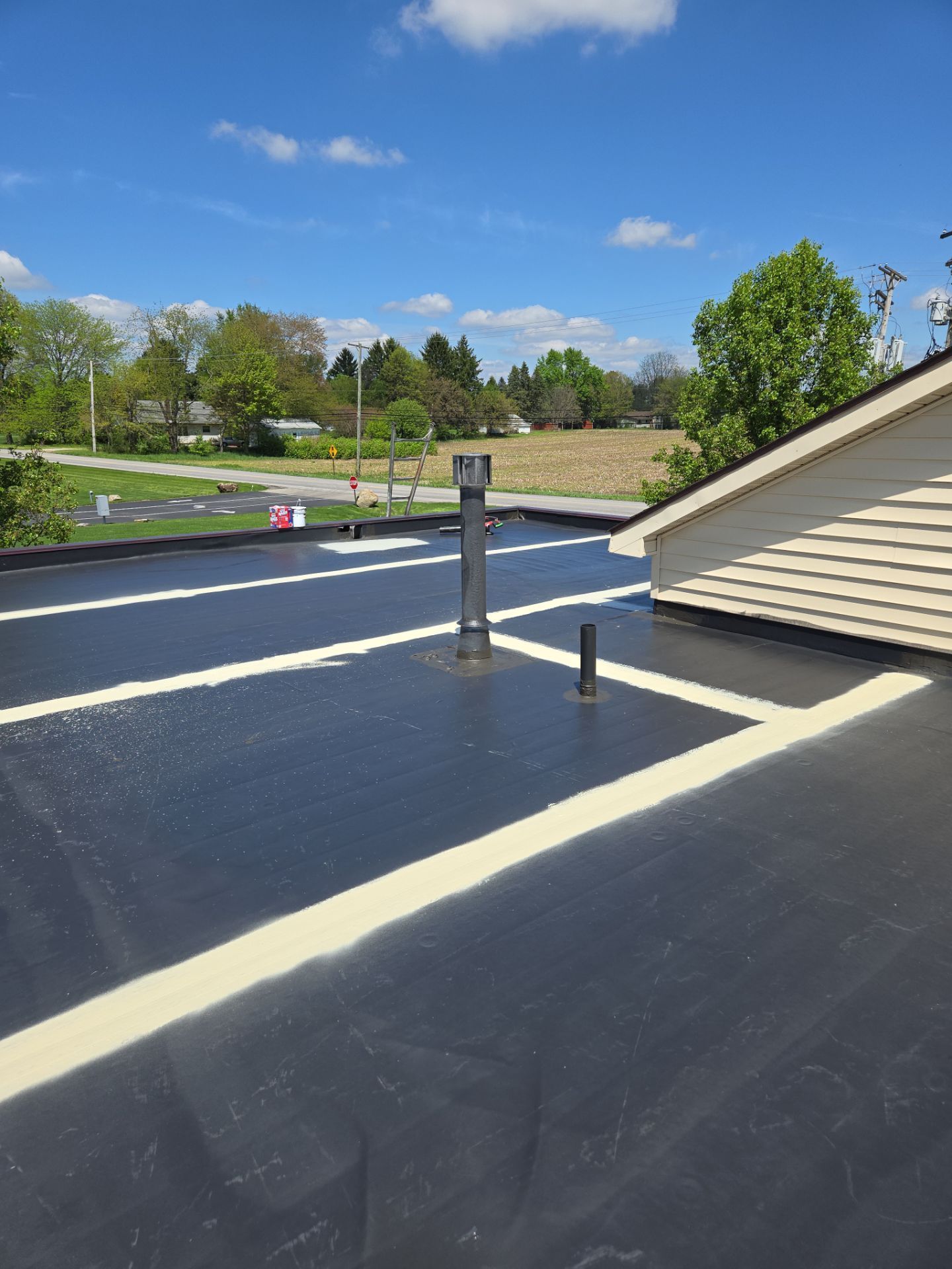 Black flat roof with white lines, a metal vent pipe, and a residential area in the background under a blue sky.