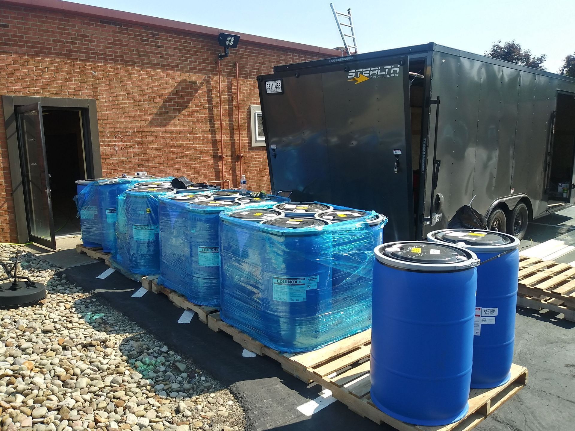 Blue barrels and wrapped containers on pallets outside a brick building next to a trailer.