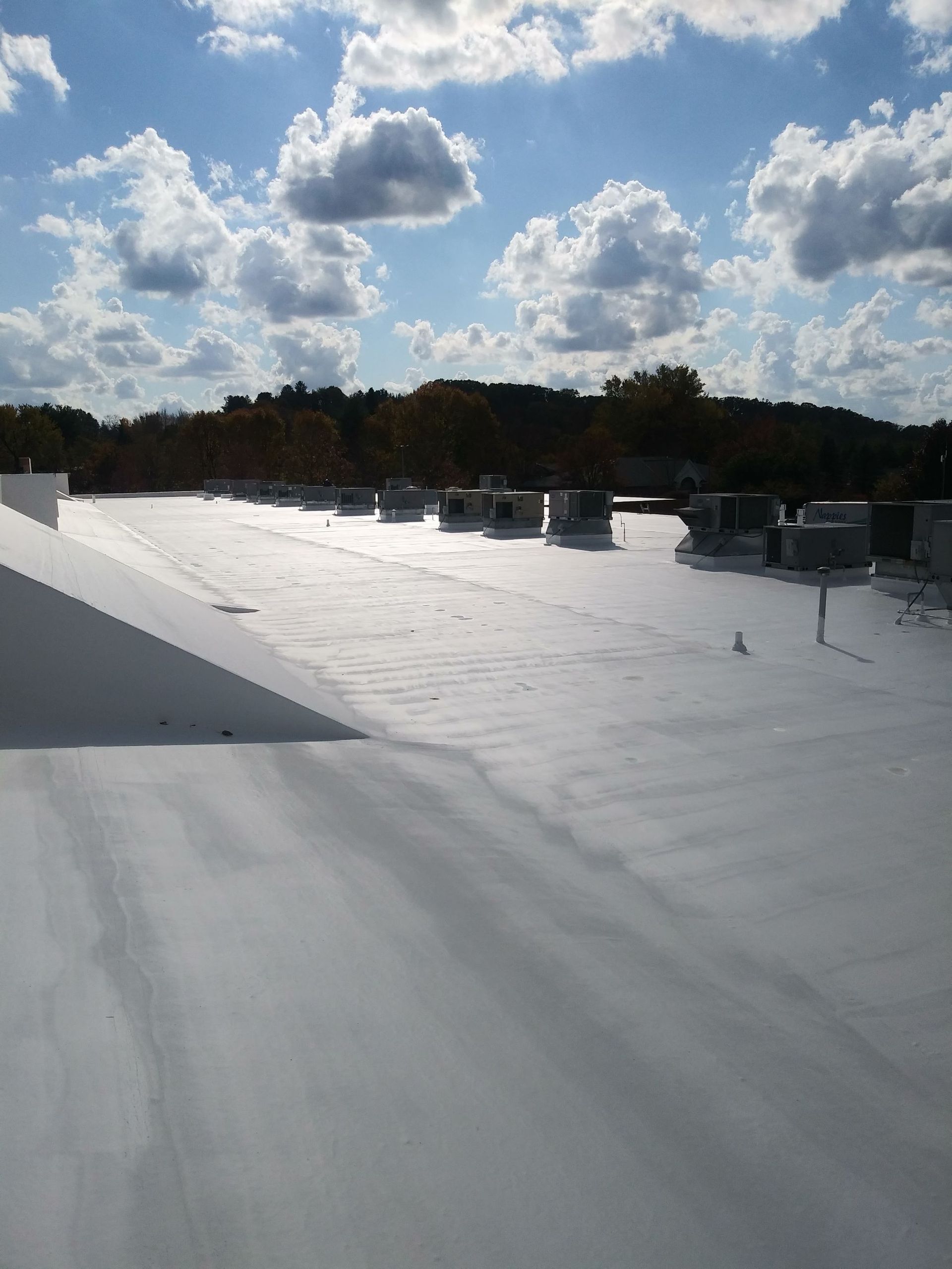 A white, flat commercial rooftop under a partly cloudy sky with HVAC units and trees in the background.