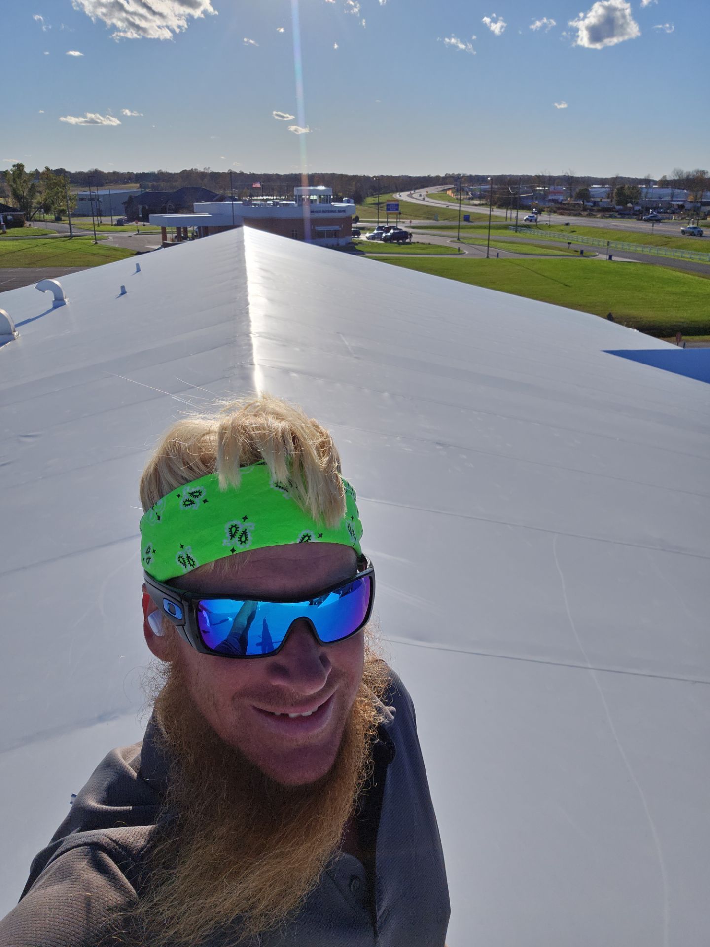 Man on a white roof wearing sunglasses and a green bandana, smiling, with a building and sunny sky in the background.