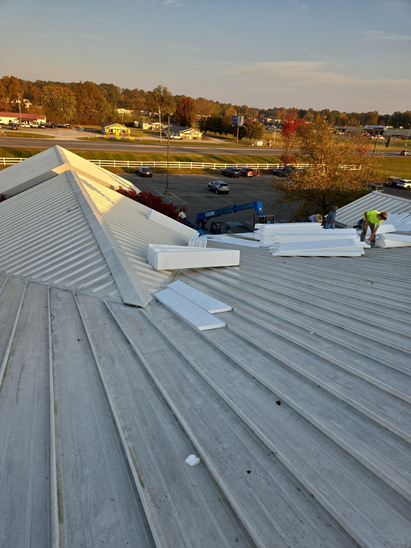 Workers installing insulation panels on a metal roof with a town in the background at dusk.