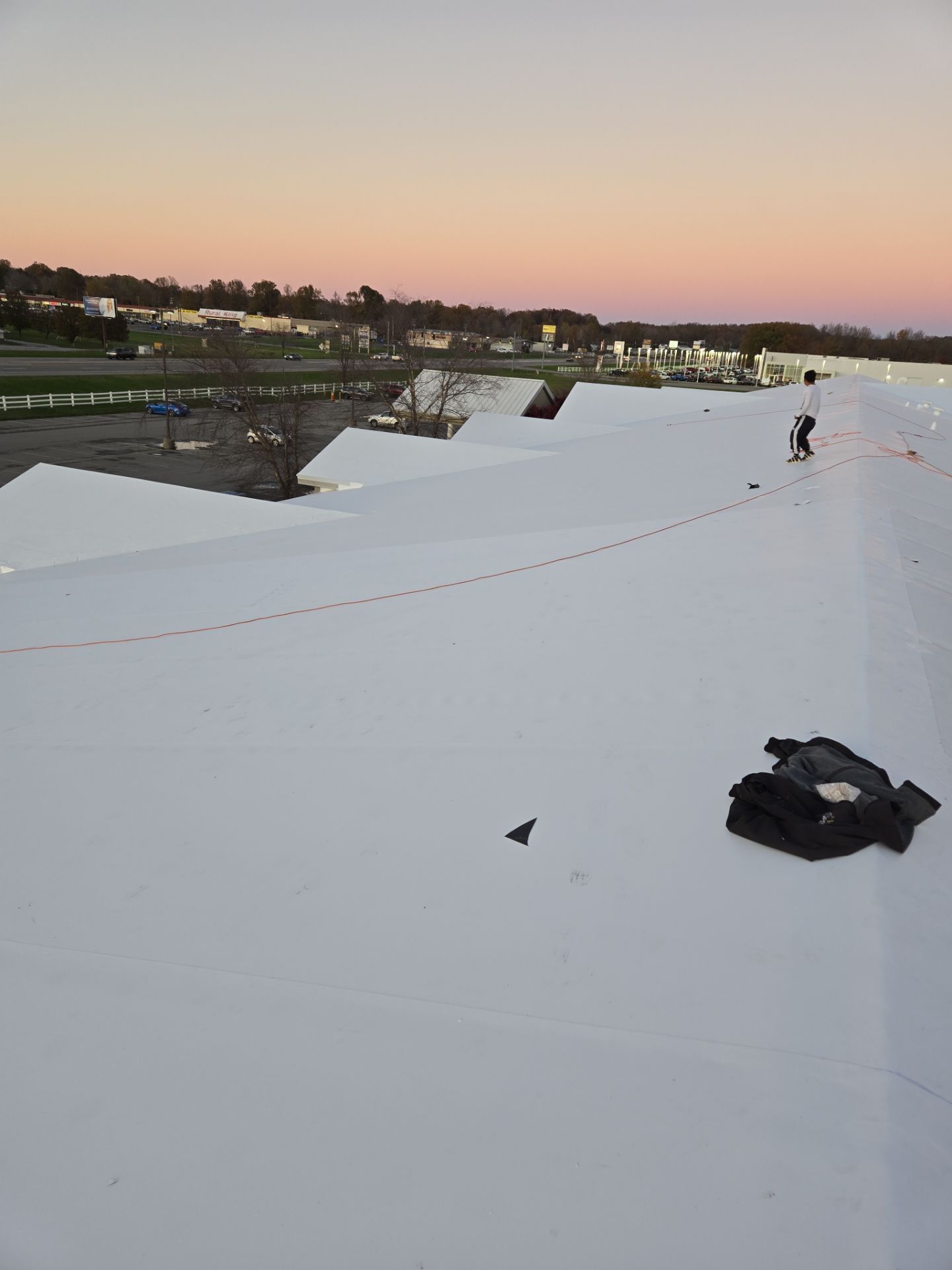 Workers on a white commercial roof at dusk. Pink and blue sky; a pile of materials in the foreground.