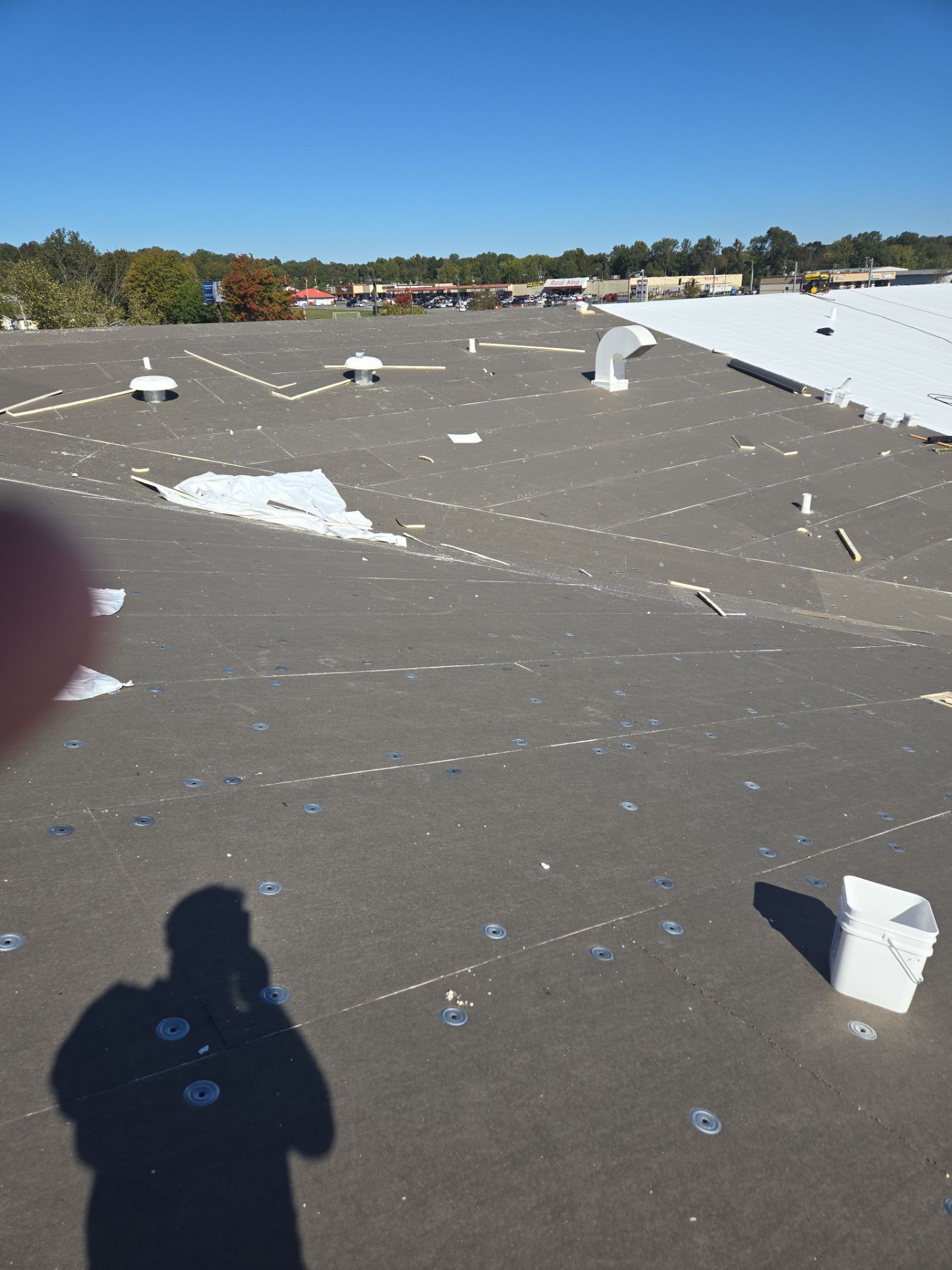 A person on a dark roof with damage. Visible vents, blue sky, and a distant commercial building.