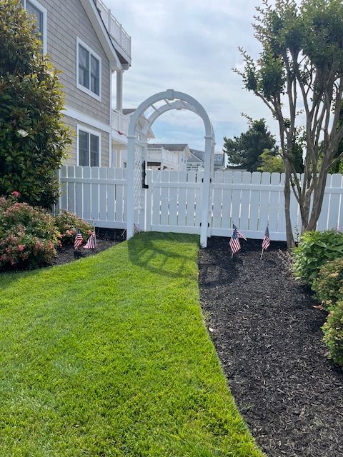 A white fence surrounds a lush green lawn in front of a house that has under deck