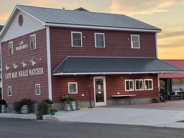 Red two-story building, Cape May Whale Watcher. Grey roof, windows, and entrance with a table outside.