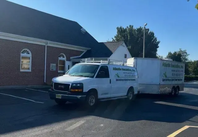 A white van towing a trailer parked in front of a building. The van has business logos on the side and a roof rack.